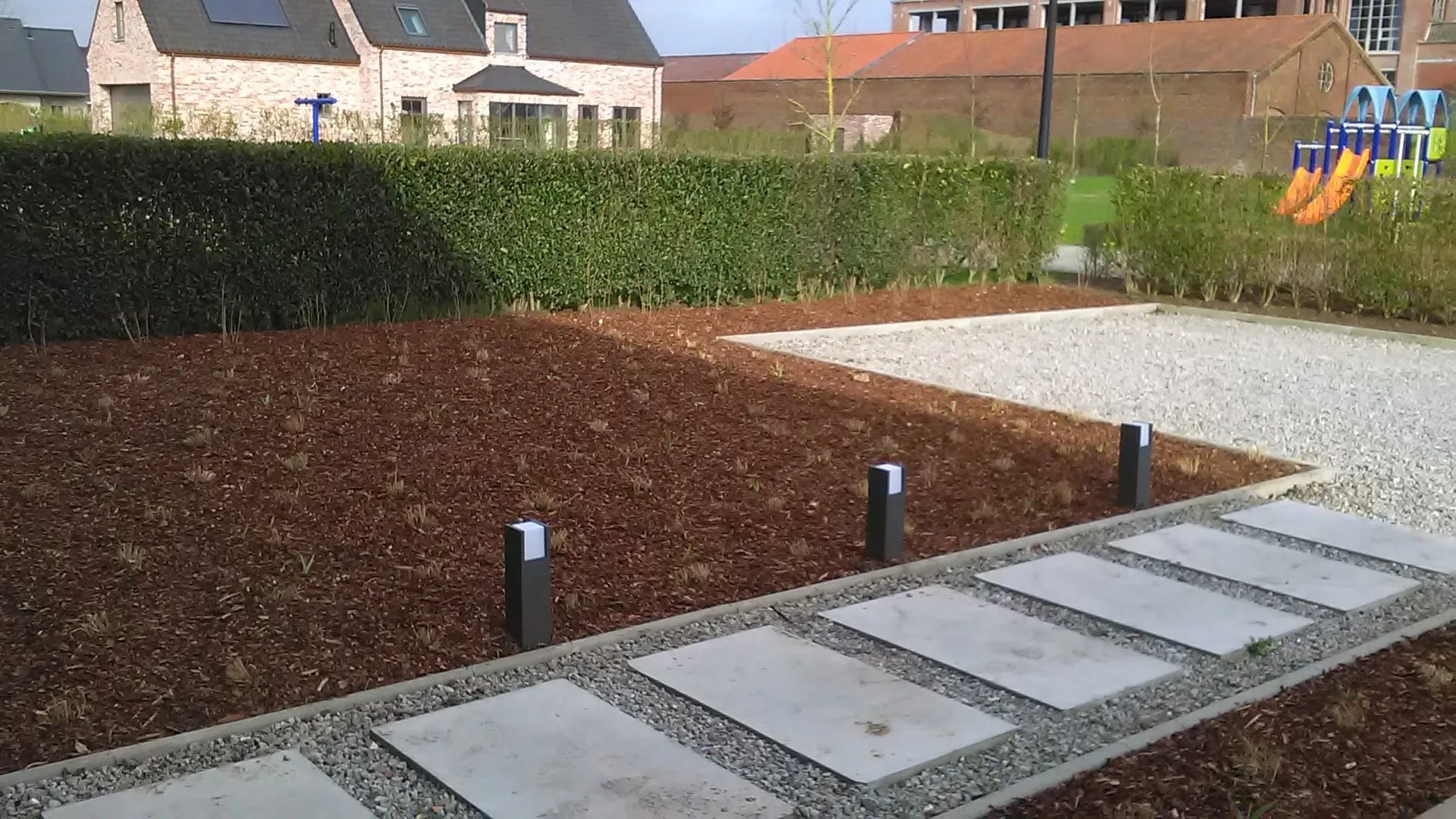 Voortuin met een plantvak in schors, betonnen staptegels in grind en kleine tuinlampen