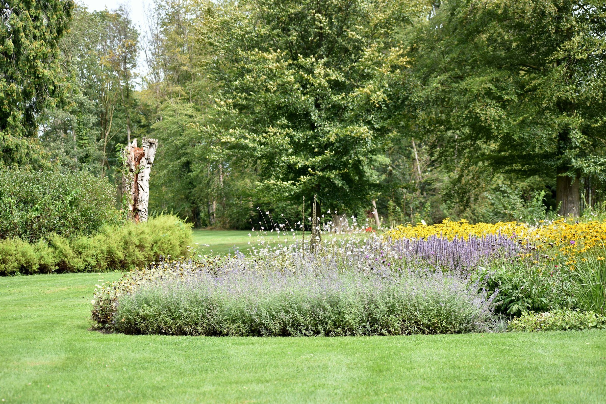 Verzorgde tuin met strak gemaaid gazon, een ronde border met vaste planten en hoge oude bomen